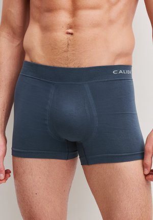 Torace maschile che indossa boxer blu con una fascia in vita visibile etichettata "CALIDA" su uno sfondo neutro.
