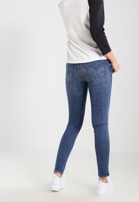 Persona vistiendo jeans ajustados azules, zapatillas blancas y una camiseta de manga raglán blanca con negro, de pie sobre un suelo gris claro.