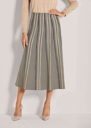 Vrouw draagt een beige en zwarte midi-rok met patroon, beige trui en beige puntige pumps, staand met één hand op haar heup.