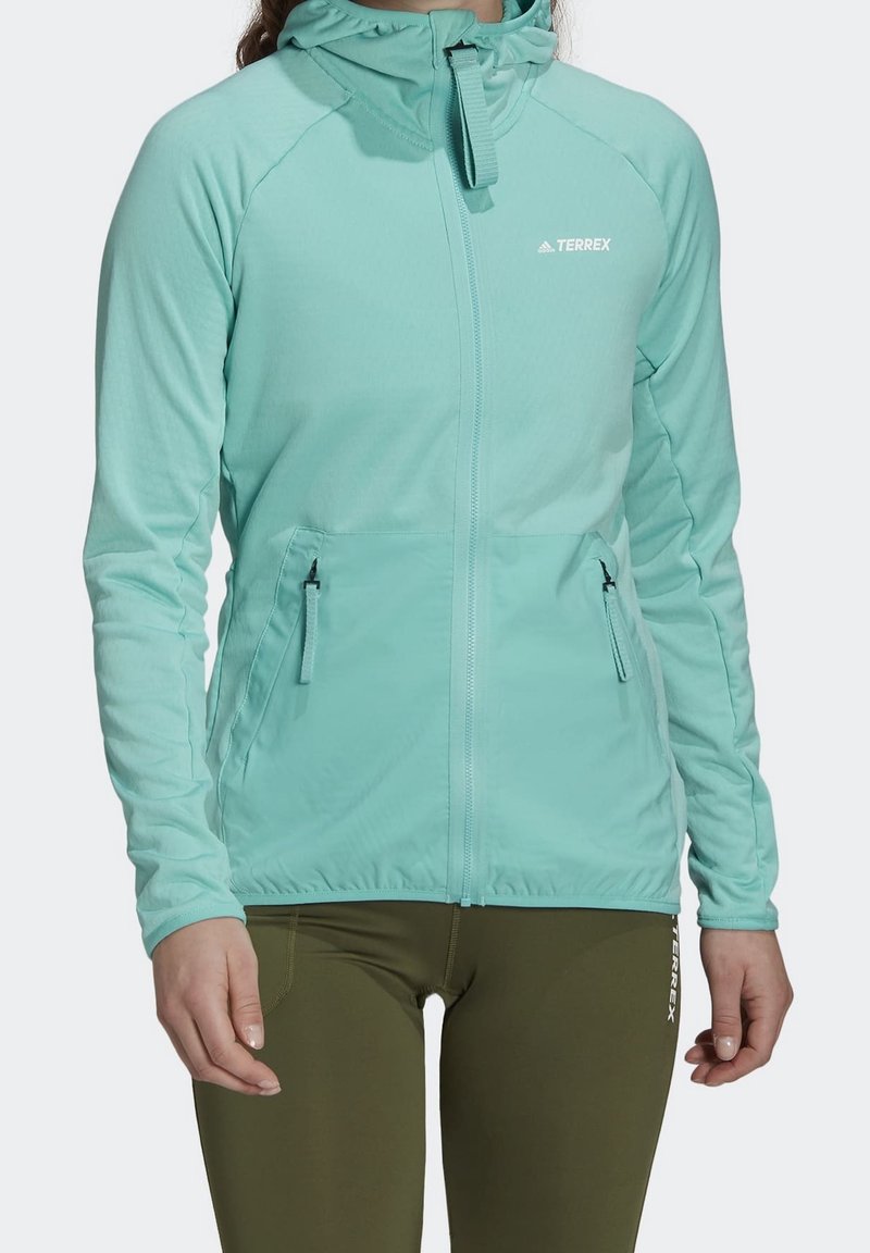 Femme portant une veste à capuche zippée bleu clair avec poches avant zippées et des leggings vert olive avec le logo Terrex.