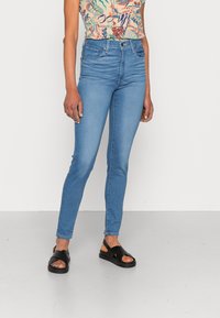 Jeans ajustados de tiro alto en un azul claro con una textura de denim suave. Combinados con sandalias planas negras con tiras entrelazadas.
