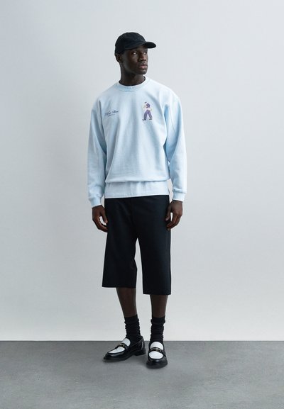 Άνδρας με ανοιχτό μπλε φούτερ με μικρό γραφικό, μαύρο σορτς μέχρι το γόνατο, μαύρο καπέλο, μαύρα και λευκά μοκασίνια με μαύρες κάλτσες, στέκεται σε γκρι πάτωμα.