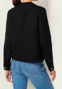 Vrouw in een gestructureerde zwarte jas met lange mouwen, voorzien van gouden knoopdetails op de manchetten, gecombineerd met hooggelegen blauwe denim jeans.