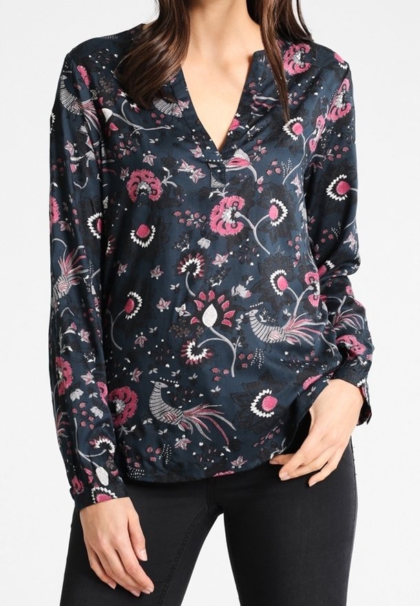 Femme portant un chemisier sombre à manches longues avec des motifs floraux et d'oiseaux roses et blancs, associé à un pantalon noir.