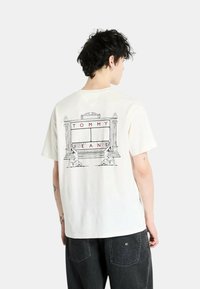 Camiseta de algodón blanca con un diseño gráfico en la parte posterior que incluye el texto "TOMMY" en rojo, rodeado de figuras ilustradas y elementos arquitectónicos.