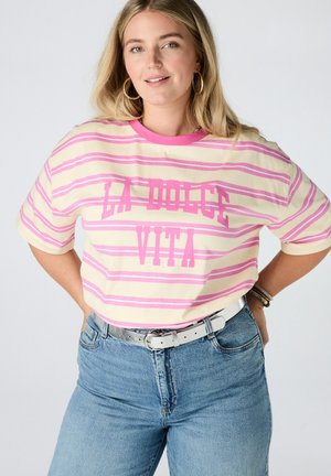 Femme aux longs cheveux blonds porte une chemise rayée rose et crème avec le texte "LA DOLCE VITA", un jean bleu, une ceinture blanche et des boucles d’oreilles en anneau.