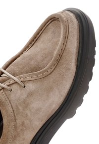 Chaussure en daim beige avec un bout arrondi, des coutures décoratives, une semelle plate en caoutchouc noir et des lacets gris clair. Texture lisse, design décontracté.