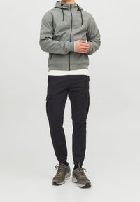 Grå zip-up hoodie med vit ribbad nederkant, tillsammans med svarta cargobyxor med sidofickor och elastiska muddar; gråa träningsskor.