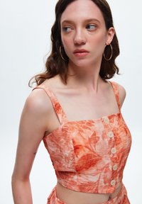 Korte top in een oranje bloemmotief met dunne bandjes, vierkante halslijn en knopen aan de voorkant. De stof lijkt lichtgewicht met een gestructureerde afwerking.