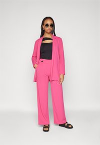 Roze blazer en wijde broek, gemaakt van een gladde stof. Zwarte top met een uitsnijding. Accessoires zijn ronde zonnebrillen en platte sandalen.