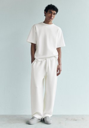 Jeune homme aux cheveux bouclés se tenant contre un mur bleu pâle, portant un t-shirt blanc ample, un pantalon large assorti et des baskets grises. Mains dans les poches.