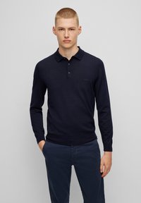 Navyblaues Langarm-Polohemd mit geripptem Material, dreiknöpfigem Verschluss und einem dezenten Logo auf der linken Brust. Getragen mit dunklen Hosen.