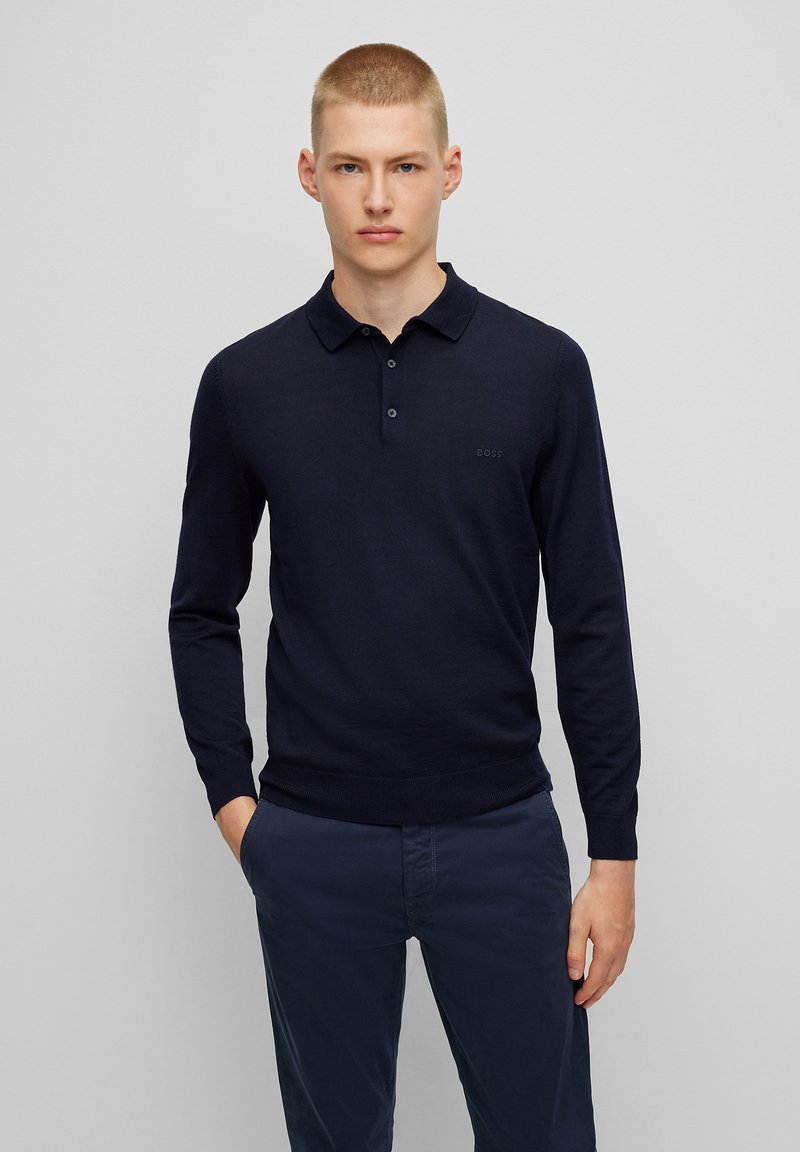 Navyblaues Langarm-Polohemd mit geripptem Material, dreiknöpfigem Verschluss und einem dezenten Logo auf der linken Brust. Getragen mit dunklen Hosen.
