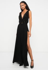 Mulher vestindo um vestido preto sem mangas, com um corpete em V profundo e com babados, e uma fenda alta, acompanhado de saltos pretos com tiras no tornozelo.