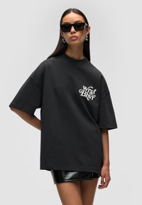 T-shirt noir oversize en coton avec des manches courtes, arborant un texte imprimé blanc. Assorti à une jupe mini noire brillante.