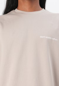 Lys beige T-shirt i blød, glat stof med rund hals og hvid tekst "don’t waste culture" trykt på venstre bryst.