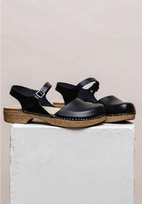 Zwarte lederen slingback klompen met houten zolen. Voorzien van een gespsluiting, ronde neus en zichtbare stiksels langs de zool.
