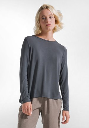 Jeune personne aux cheveux blonds courts portant un haut à manches longues gris foncé et un pantalon marron clair sur fond blanc.