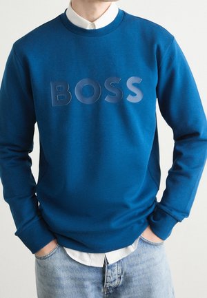 Osoba nosząca niebieską bluzę z napisem "BOSS" dużymi literami, biała koszula pod spodem oraz jasnoniebieskie dżinsy z rękami w kieszeniach.