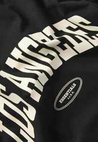 T-shirt in tessuto nero con grande testo stilizzato bianco "LOS ANGELES" e un piccolo logo ovale "ESSENTIALS J.J.X.X." in basso.