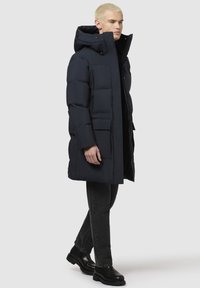 Tengerészeti sötétkék leleződzseki leválasztható kapucnival, cipzáras elülső résszel, oldalsó zsebekkel és textúrált felülettel. Fekete farmernadrág és vastag cipők fölött viselve.