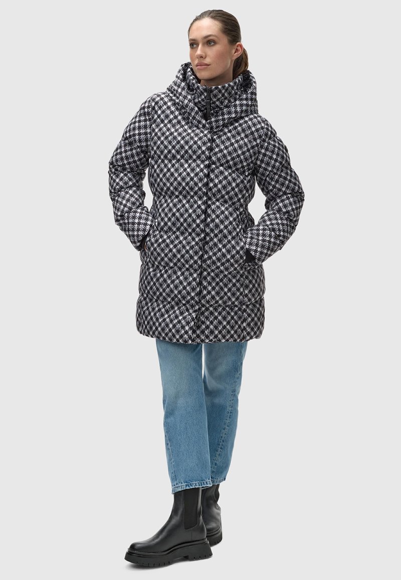 Chaqueta acolchada con patrón en blanco y negro, con cuello alto, cierre de cremallera y bolsillos, combinada con vaqueros azul claro y botas negras de tobillo.