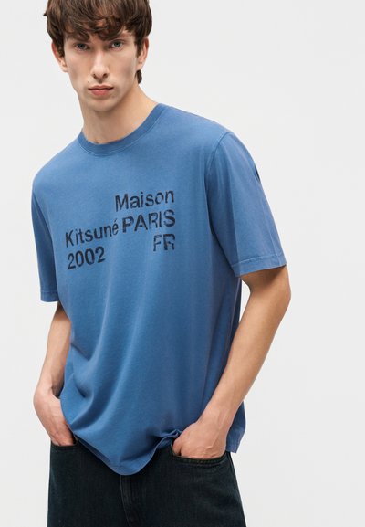 Νεαρός άνδρας που φοράει μπλε μπλουζάκι Maison Kitsuné Paris 2002 FR με τυπωμένο σχέδιο, με τα χέρια στις τσέπες σκούρων παντελονιών, κοιτάζοντας μπροστά.