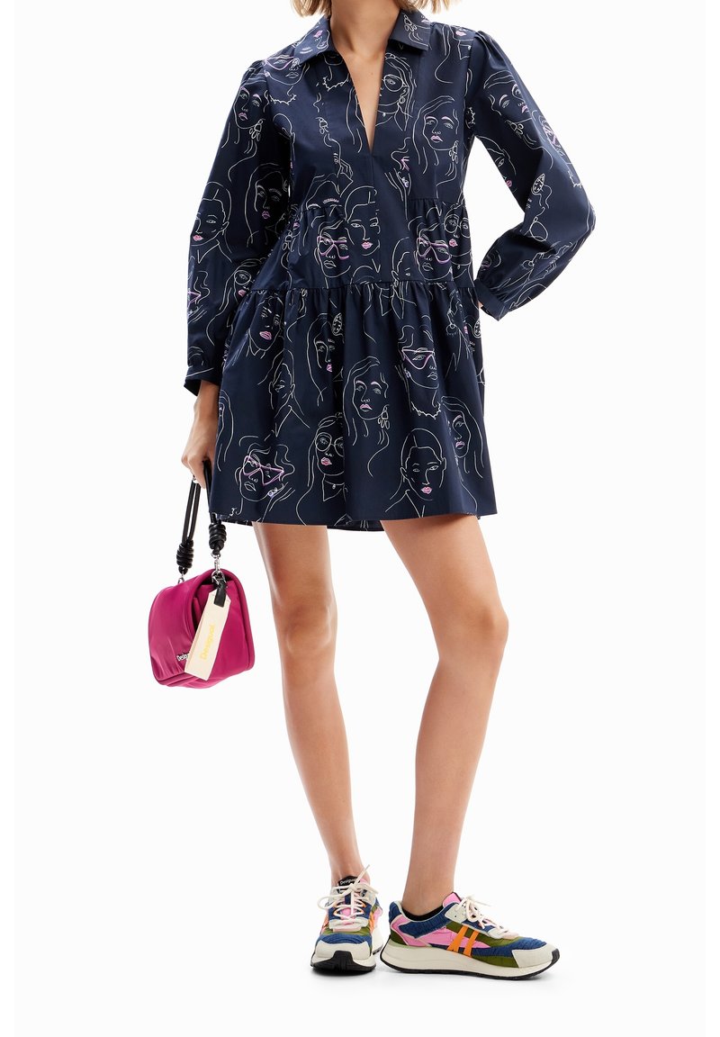 Femme portant une robe bleu marine avec des imprimés abstraits de visages, tenant un petit sac à main rose, et des baskets multicolores sur fond blanc.