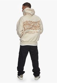 Kremfarget hettegenser med brun tekst på baksiden, "DROPSIZE CLUB / EST. 2020," kombinert med svarte cargo-bukser og grå sneakers.