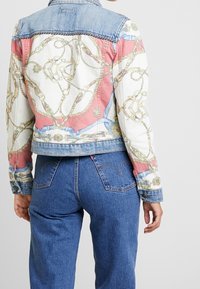 Veste en denim avec un dos imprimé coloré orné de chaînes et de motifs décoratifs, col bleu clair contrastant et poignets boutonnés.