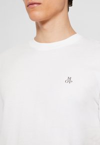 T-shirt bianca in cotone con scollatura rotonda e maniche corte. Presenta un piccolo logo nero "M OP" ricamato nell'area sinistra del petto.