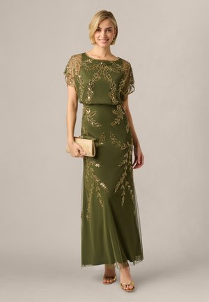 Donna che indossa un abito lungo verde oliva con ricamo di foglie dorate, tiene una clutch dorata e indossa tacchi aperti dorati, sorridendo alla fotocamera.