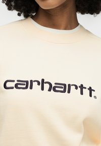 Κοντινό πλάνο ενός ατόμου που φοράει μια κρεμ φούτερ με μαύρα κεντημένα γράμματα «carhartt» στο στήθος.