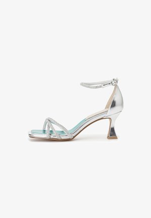 Sandale argentée à fines brides avec un petit talon bloc, des brides ornées et une semelle intérieure turquoise. Dispose d'une bride à la cheville avec une fermeture à boucle.