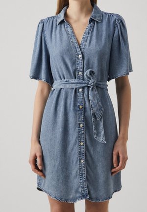 Mujer con un vestido camisero corto de mezclilla azul con cinturón y botones en la parte delantera, sobre un fondo liso.