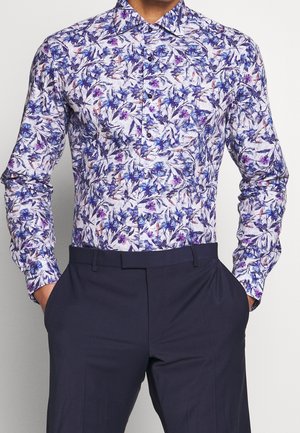 Camicia a maniche lunghe con bottoni e motivo floreale blu e viola, indossata con pantaloni blu scuro, mani in tasca, volto non visibile.