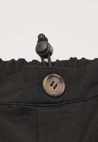 Tissu noir avec une finition texturée, doté d'une ceinture élastique, d'un bouton marron à deux trous et d'un cordon noir avec un bouton-pression pour l'ajustement.