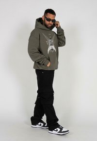 Hanorac verde măslinie cu imprimeu grafic, buzunar frontal, pantaloni negri de stil cargo și adidași alb-negru. Textură de țesătură netedă.