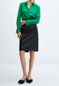 Blouse verte boutonnée avec manches longues, jupe crayon en cuir noir, et chaussures noires avec accents de nœuds. Design simple et ajusté.