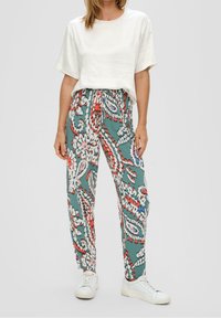 Luźne zielone spodnie w wzory z akcentami czerwieni, bieli i niebieskiego; dobrane do kremowego, krótkiego rękawa i białych sneakersów.