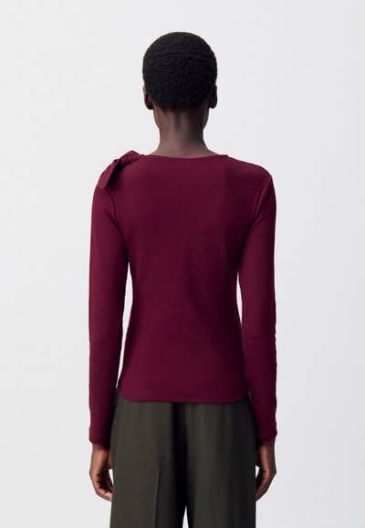 Top bordeaux a maniche lunghe con una texture morbida, dotato di un dettaglio a fiocco sulla spalla sinistra e un collo rotondo.