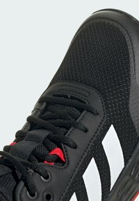 Zapatilla deportiva negra con parte superior de malla, tres franjas blancas, acentos rojos y cordones negros. Suela de goma texturizada y cuello acolchado.
