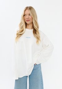 Witte lange mouwen shirt met blauwe "MIRONS" tekst. Losse pasvorm; zachte, lichte stof. Gecombineerd met lichtblauwe spijkerbroek, handen in de zakken.