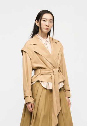 Kvinde med glat sort hår iført en beige trenchcoat med bælte over en stribet skjorte og plisseret tan nederdel, står foran en ensfarvet baggrund.