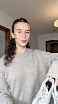 Sivi pleteni džemper s rebrastim ovratnikom, lagano širokim krojem, nošen od strane osobe koja drži bijele tenisice s crnim i sivim detaljima.