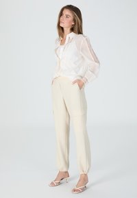 Weiße, transparente Bluse mit Spitzenbesatz, kombiniert mit beigen Cargo-Hosen mit Seitentaschen. Das Model trägt schicke Sandalen mit Riemchen und steht vor einem hellen Hintergrund.