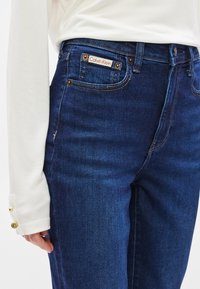 Σκούρο μπλε τζιν με ψηλή μέση, παραδοσιακό σχέδιο με πέντε τσέπες, ορατές ραφές και ετικέτα με το λογότυπο Calvin Klein στην πίσω τσέπη.