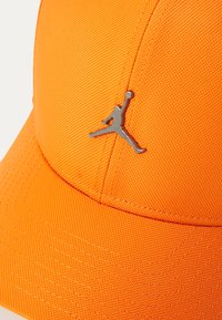 Orange baseballcap lavet af struktureret stof, med et metallisk sølv Jumpman-logo på forsiden.
