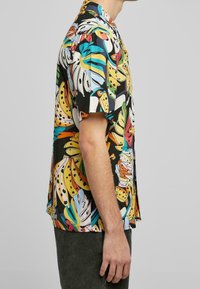 Chemise à manches courtes avec une base noire ornée de motifs colorés de bananes et de feuilles. Confectionnée en tissu léger, elle est conçue pour une coupe décontractée.