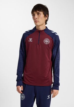 Mann trägt burgunderrotes und marineblaues Sporttop und Hose mit Logos des Dänischen Fußballverbands, steht vor weißem Hintergrund.
